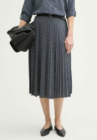 Schwarz-blauer plissierter Rock mit geometrischem Muster, kombiniert mit einem taillierten schwarzen Gürtel und einer schwarzen Schultertasche. Getragen mit schwarzen flachen Schuhen.