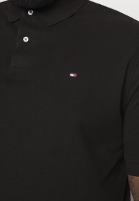Černé polo tričko s texturovanou tkaninou, s léga na tři knoflíky a malým barevným logem na levé hrudi.