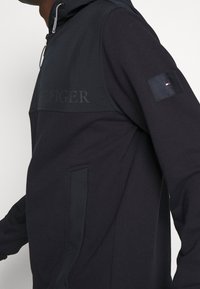 Sudadera con capucha azul marino con un logo tonal en el pecho, bolsillo lateral y parche con bandera en la manga. Textura suave con diseño deportivo.