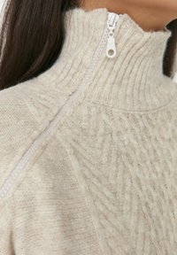 Texturierter, cremefarbener Pullover mit geripptem Kragen und halblangem Reißverschluss, der ein gestricktes Muster und eine weiche, flauschige Textur aufweist.
