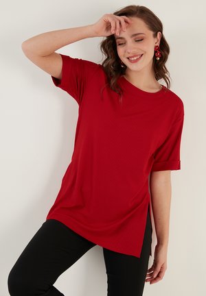 Maglietta rossa a coste con scollo rotondo, maniche corte, spacchi laterali e vestibilità rilassata, abbinata a pantaloni neri.