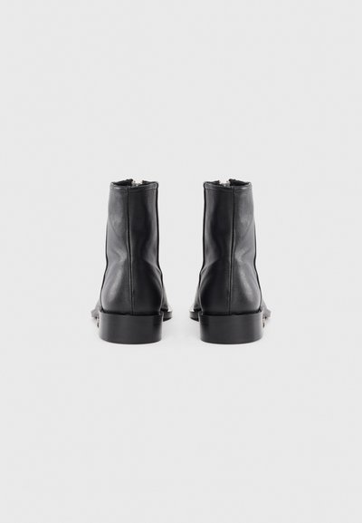 Botas de piel negras hasta el tobillo con una textura suave, que presentan un tacón bajo y robusto, y un diseño minimalista. Cremalleras en la parte trasera para facilitar su uso.