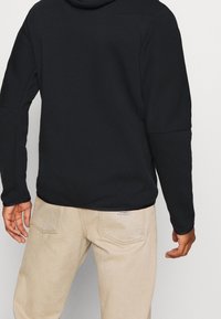 Man som bär en svart långärmad tröja och beige byxor, sedd bakifrån, med avslappnade händer vid sidorna.