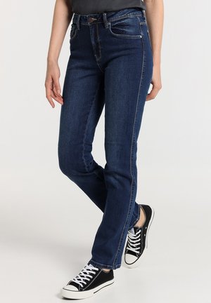 Vaqueros de denim azul oscuro con corte ajustado, diseño de cinco bolsillos, ansas para cinturón y cierre de botón, combinados con zapatillas de lona negras.