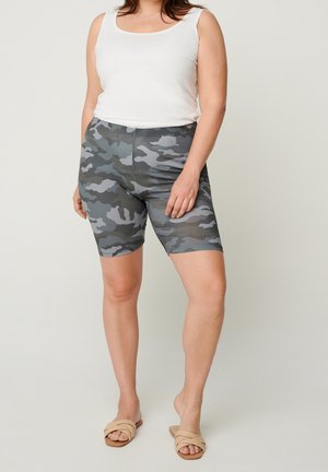 Kvinde iført grå biker-shorts med camouflage-mønster, hvid ærmeløs top og beige slide-sandaler, stående mod en neutral baggrund.