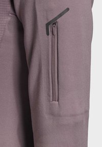 Sudadera de color púrpura claro con tejido texturizado. Cuenta con un bolsillo con cremallera en la manga izquierda y un acento de diseño negro encima del bolsillo.