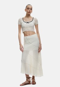 Spitzen-Crop-Top mit kurzen Ärmeln, weiß mit schwarzem Rand und Schleifendetail; geschichteter, halbtransparenter weißer Rock mit strukturiertem Zickzackmuster.