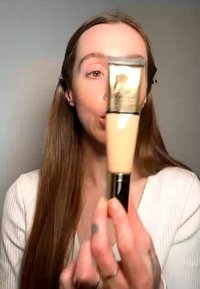 Kvinde med langt brunt hår holder et rør med beige flydende foundation tæt på sit ansigt mod en ensfarvet baggrund.