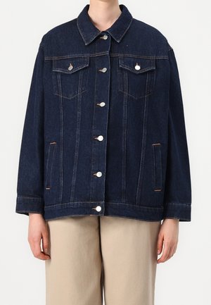 Chaqueta vaquera de mezclilla azul oscuro con cierre de botones, bolsillos delanteros con solapa y mangas largas, llevada sobre pantalones beige.