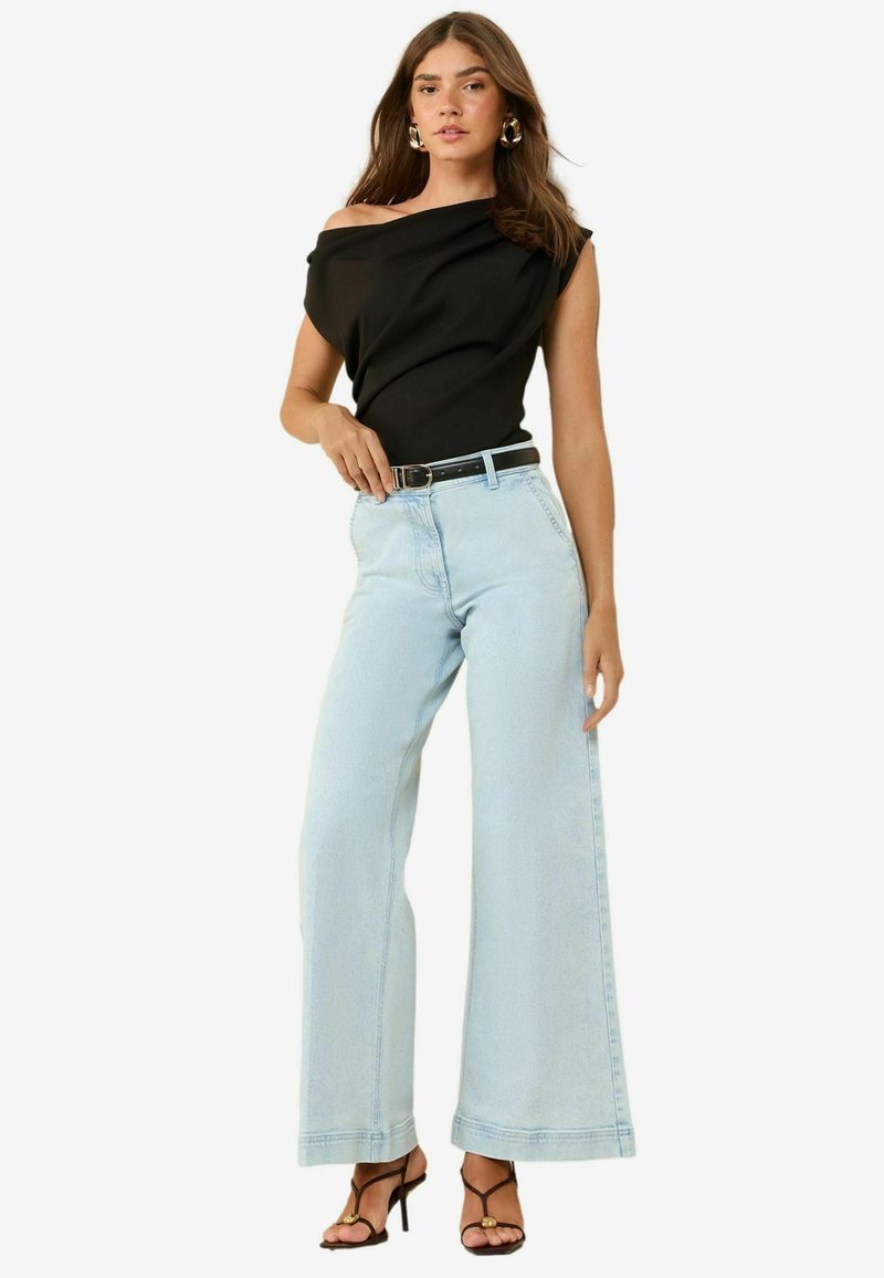 Top nero scoperto sulle spalle, jeans baggy azzurri, e cintura nera. Modella indossa tacchi neri a strappo con una texture liscia.