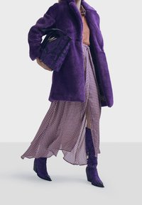 Abrigo de piel sintética morado, vestido estampado en púrpura y beige, botas moradas con estampado y un bolso negro con flores, creando un atuendo atrevido y coordinado.