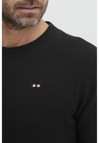Juodas megztinis su apvaliu kaklu ir mažą, raudonai, baltai ir mėlynai siuvinėtą logotipą ant krūtinės.