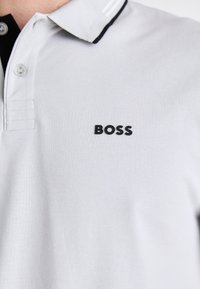 Biely polo tričko s textúrovanou tkaninou, s čiernym logom "BOSS" na ľavej strane a kontrastným čiernym akcentom na golieri.
