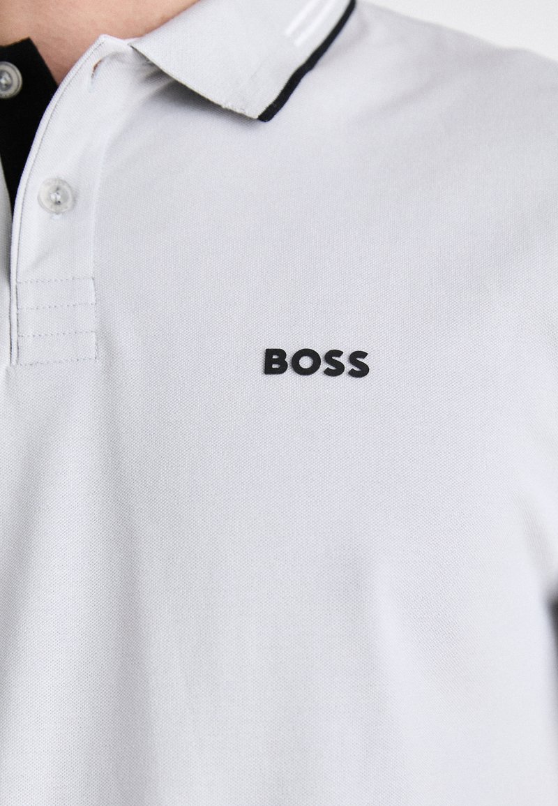 Polo blanc en tissu texturé, avec un logo noir "BOSS" sur le côté gauche et un accent noir contrastant sur le col.