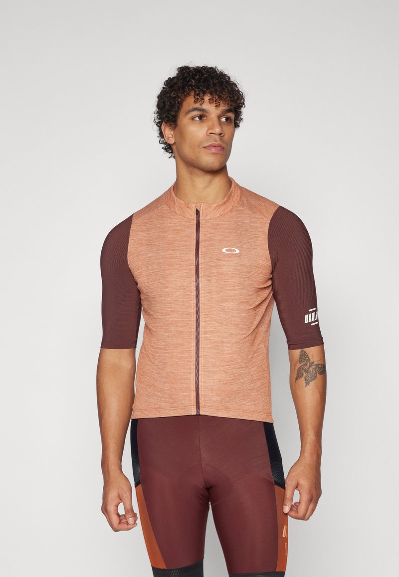 Cyklistický dres s krátkým rukávem v odstínech oranžové a hnědé, strukturovaná látka, přední zapínání na zip, logo na hrudi a rukávu.