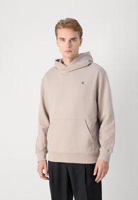 Beige pamut kapucnis pulóver kenguruzsebbel, bordás mandzsettákkal és egy diszkrét logóval a mellkasán. Állítható kapucnival rendelkezik.