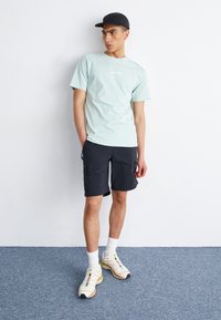 Lys blå t-skjorte, svarte cargo-shorts, hvite og multicolor sneakers, svart caps, står på et blått teppe mot en hvit vegg.