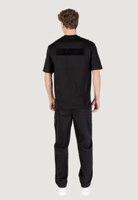 La camiseta negra de manga corta presenta una etiqueta rectangular en la parte posterior con un logotipo en relieve. Se combina con pantalones negros holgados y zapatillas blancas.