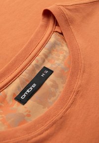 Ombre oranžové tričko s kulatým výstřihem. Vnitřní strana má motiv maskáče v broskvové a světle šedé barvě. Viditelná velikostní etiketa.