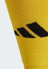 Żółta skarpetka piłkarska wykonana z teksturowanej tkaniny, z wyraźnym czarnym wzorem trzech pasków blisko góry.