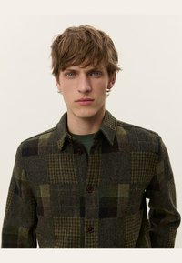 Jachetă căptușită verde cu carouri, cu guler, având texturi și modele variate. Accente cu nasturi maroni. Modelul poartă o cămașă verde dedesubt.