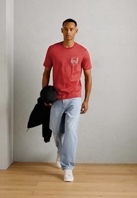 Rdeča bombažna majica s skritim žepom na prsih, v kombinaciji s svetlo modrimi kavbojkami in belimi tenisicami, model drži črno jakno.