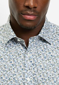 Florales Hemd zum Knöpfen in Hellblau mit Blattmustern in Grün und Beige. Klassischer Kragen und weiße Knöpfe. Baumwollmaterial.