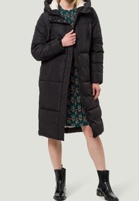 Abrigo acolchado negro con capucha, que presenta un diseño acolchado y cierre frontal. Llevado sobre un vestido floral, combinado con botas de tobillo negras brillantes.