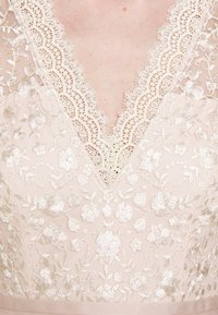 Gros plan d'un vêtement en dentelle beige avec broderie florale et bords festonnés autour d'un décolleté en V sur une peau claire.