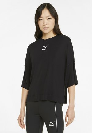 Maglietta nera oversized con maniche corte, caratterizzata da un logo Puma bianco al centro. Il materiale sembra soffice e liscio.