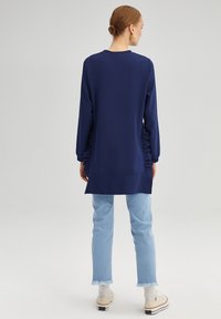 Maglia a maniche lunghe blu navy con dettaglio arricciato sui lati, scollatura rotonda e vestibilità comoda. Abbinata a jeans azzurri e sneakers bianche.