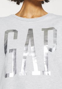 Camiseta de algodón gris con grandes letras en bloque metálicas plateadas que dicen "GAP". Cuello redondo y mangas cortas. Textura suave.