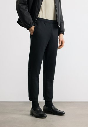 Άνδρας που φοράει μαύρο παντελόνι τζόγκινγκ, μαύρα δερμάτινα μοκασίνια, μαύρο μπουφάν και ανοιχτό μπεζ πουκάμισο, στέκεται με το ένα χέρι στην τσέπη.