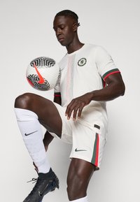 Maglia da casa del Chelsea FC, bianca con accenti rossi e verdi, caratterizzata da un logo della squadra. Il giocatore tiene in mano un pallone da calcio Nike in bianco e nero, indossando scarpini Nike.