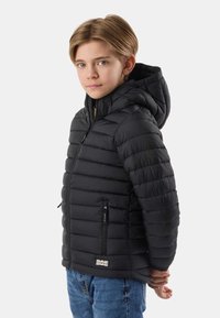 Garçon blond portant une doudoune à capuche noire et un jean bleu, debout de profil avec les mains derrière le dos sur un fond uni.