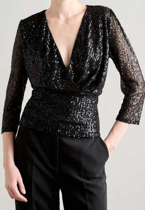 Blouse noire à sequins cache-cœur avec manches transparentes trois-quarts, associée à un pantalon noir taille haute, modèle posant avec une main dans la poche.