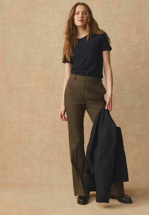 Noor naine pikkade juustega seisab, kandes musta t-särki, pruune ülikonnapükse ja musti kingi, hoides käes musta jopet beeži tausta ees.