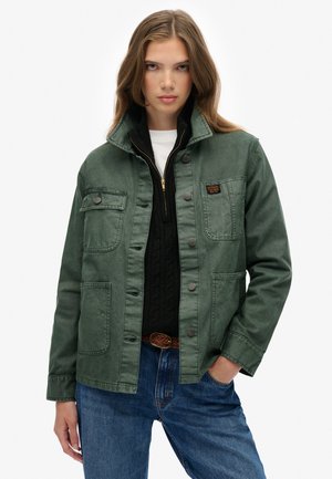 Groene jasje met hoge kraag, voorknopsluiting en twee borstzakken; gedragen over een zwart gebreid vest en blauwe spijkerbroek.
