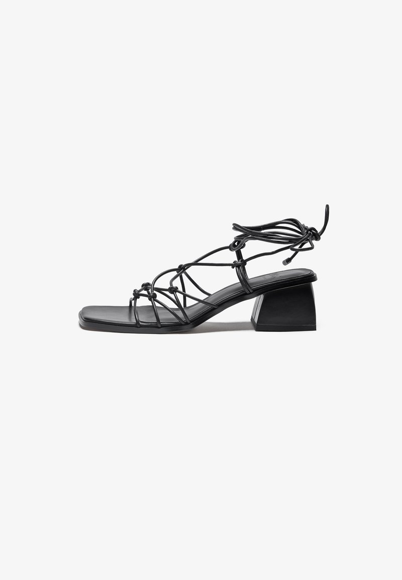 Black strappy sandal with a square toe, featuring a knotted design and a chunky, geometric heel for stability. Made of smooth material.