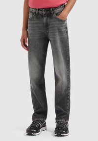 Person trägt ausgewaschene schwarze, gerade geschnittene Jeans, schwarze und weiße Sneaker sowie ein korallenrotes Oberteil und hat eine Hand in der Tasche.