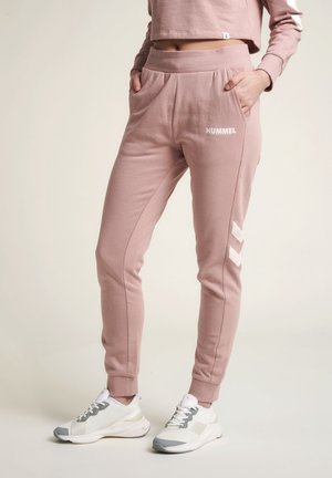 Persoon die roze Hummel trainingsbroek met zakken draagt en witte sneakers, staand tegen een effen lichte achtergrond.