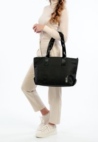 Kobieta w beżowym stroju trzyma dużą czarną torbę na ramię z krótkimi uchwytami, nosząc białe sneakersy na platformie na tle białym.