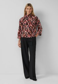 Langarmshirt mit hohem Kragen in rosa, braun und schwarz, mit einem auffälligen abstrakten Muster, kombiniert mit schwarzen weiten Hosen.