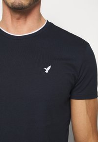 Mörkblå bomulls t-shirt med en vit ribbad krage och en liten broderad vit fågel på vänster bröst. Korta ärmar och en figursydd design.