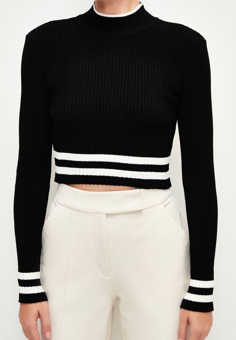 Crop top noir à côtes avec col haut et manches longues. Présente des bandes blanches horizontales aux poignets et à l'ourlet. Tissu doux et texturé.