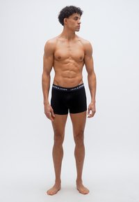 Fit young man standing barefoot, wearing black Jack & Jones boxer briefs, looking right against plain light gray background.