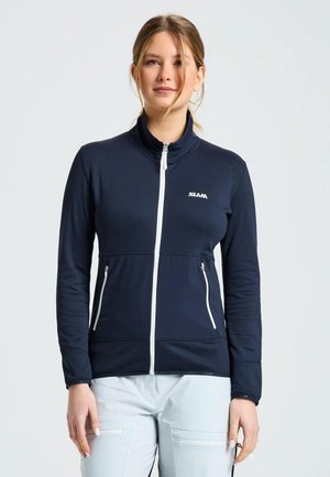 Una mujer luce una chaqueta azul marino con cremallera y ribetes blancos, junto con unos pantalones claros a juego, sobre un fondo liso.