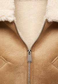 Veste couleur noisette doublée de shearling avec une texture extérieure en daim lisse, dotée d'une fermeture éclair en métal et de deux poches latérales.