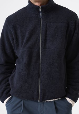 Mann trägt eine dunkelblaue Fleecejacke mit durchgehendem Reißverschluss und Brusttasche, die Hände in die Hosentaschen gesteckt.