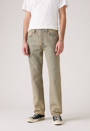 Man wearing light brown straight-leg jeans, white t-shirt, black low-top sneakers, and a silver bracelet on left wrist.